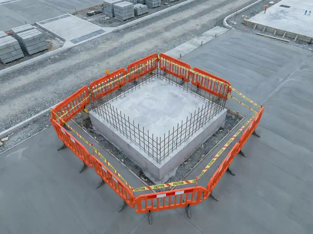Betonnen fundament hoek met wapeningsstaven, omringd door oranje veiligheidshekken op bouwplaats