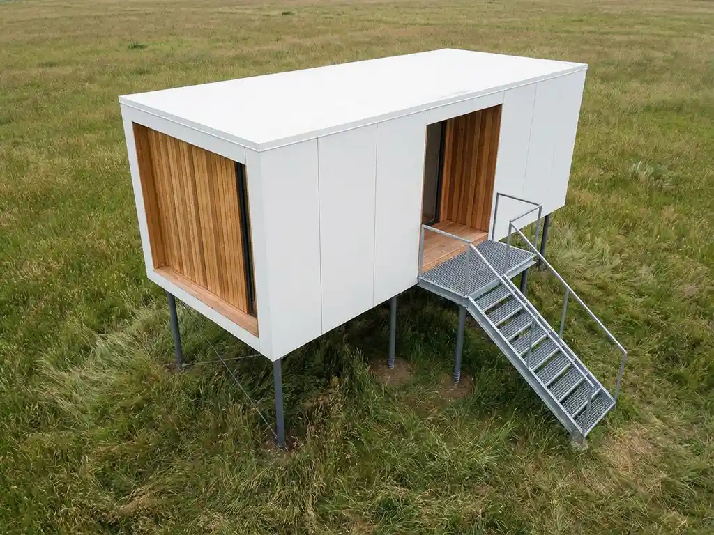 recreatiecabine-stalen-schroeffunderingen-modern-ontwerp.jpg Moderne recreatiehut op stalen schroeffunderingen boven natuurlijk gras, witte en houten gevel met geometrische lijnen