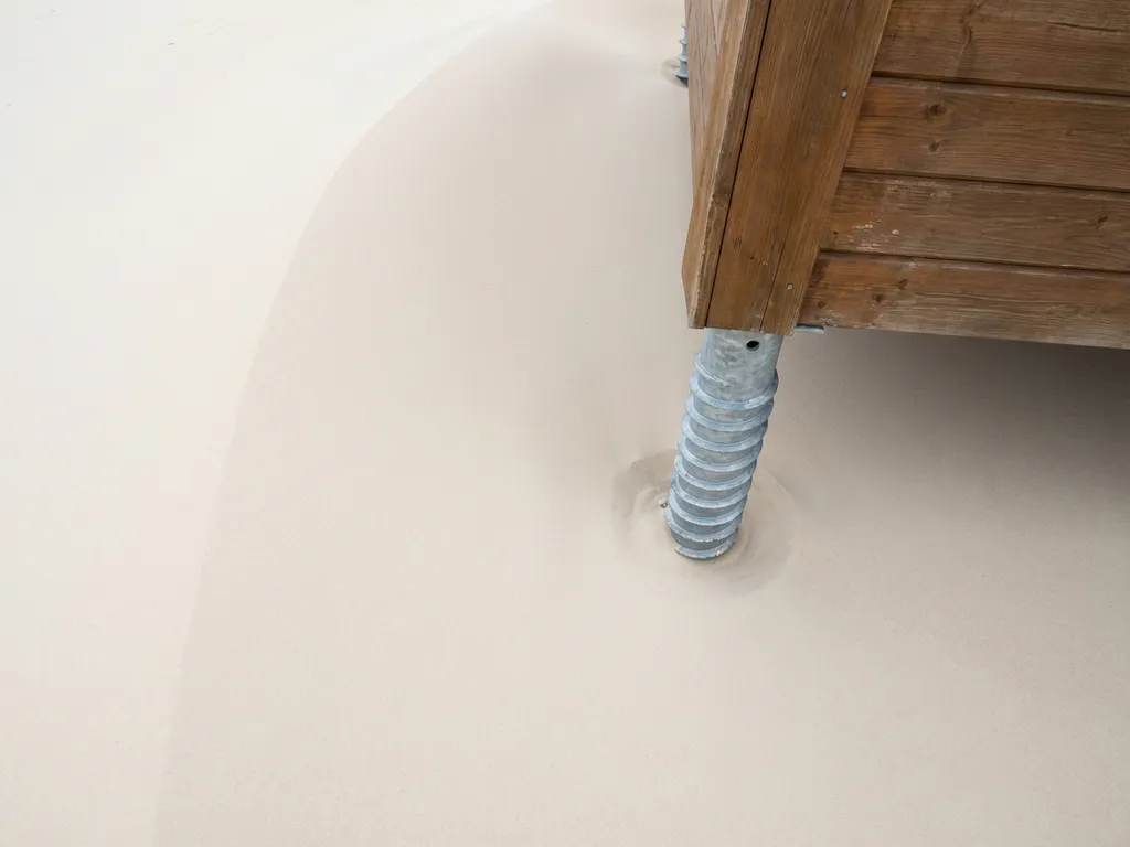 Stalen schroeffundatie naast houten recreatiecabine op zandige ondergrond, spiraalvormige draad zichtbaar