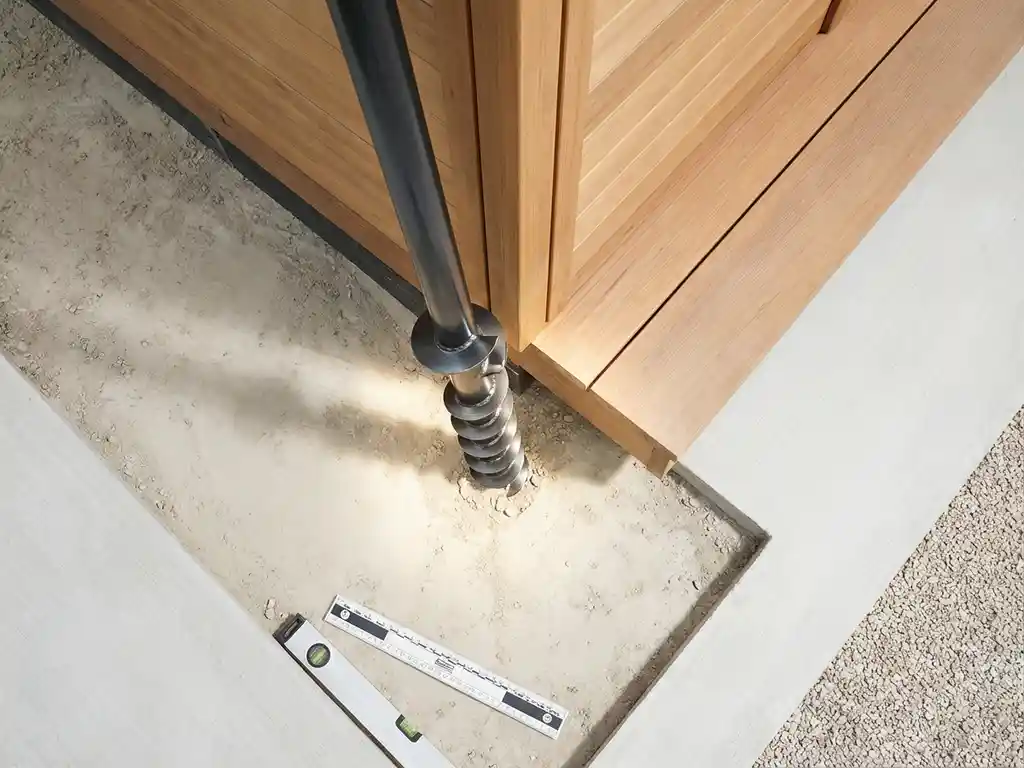 schroeffundering-installatie-tuinhuis-staal-fundatie.jpg Stalen schroeffundatie wordt geïnstalleerd naast modern houten tuinhuis, bovenaanzicht met geometrische lijnen