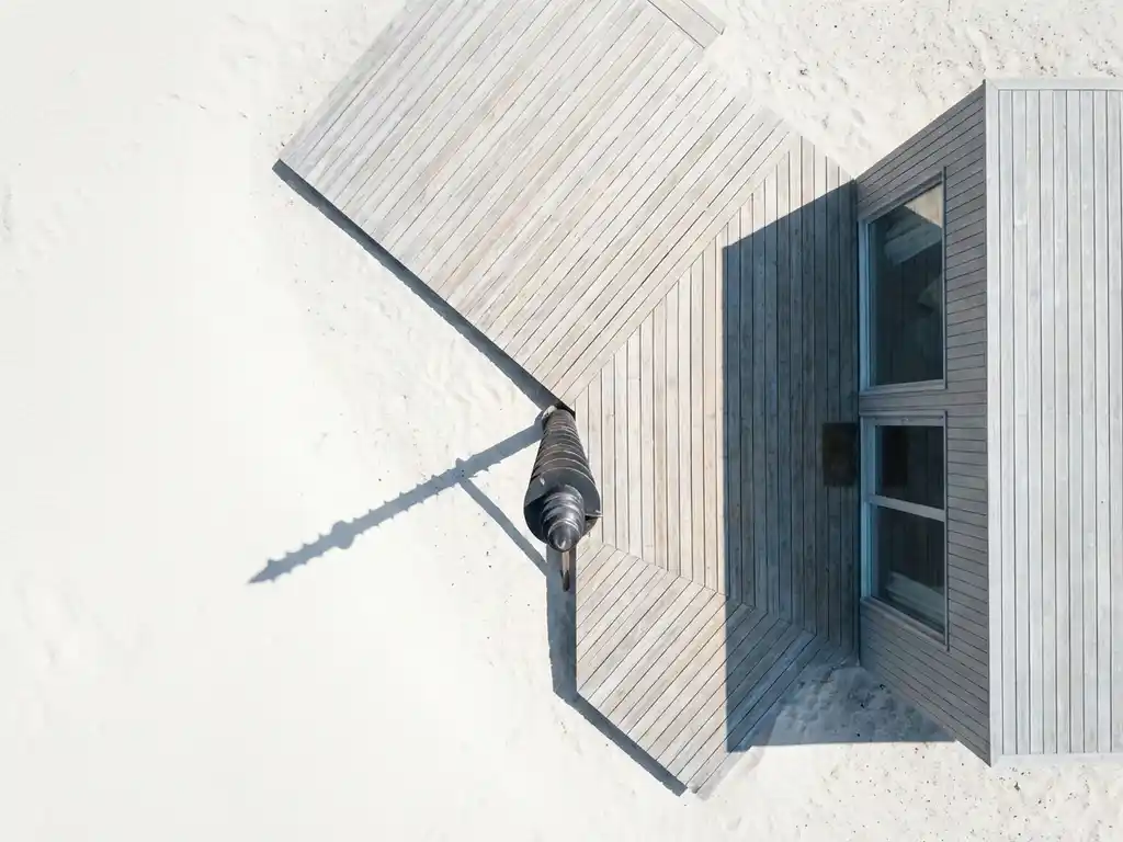 Stalen schroefpaal met spiraalschroefdraad steekt uit wit zand naast minimalistisch houten vakantiehuis met geometrisch terras