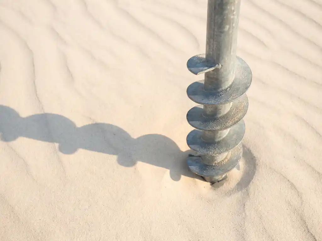 schroefpaal-gegalvaniseerd-staal-zand-installatie.jpg Gegalvaniseerde stalen schroefpaal in wit zand met helixvormige schroefdraad en geometrische schaduwen