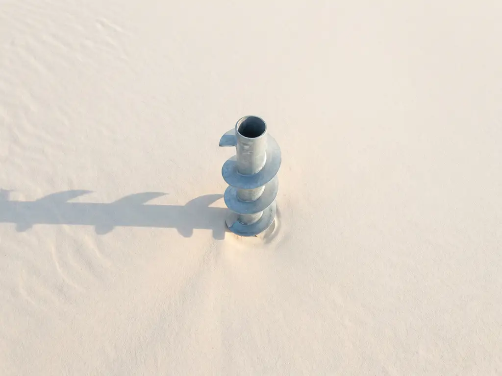 schroefpaal-gegalvaniseerd-staal-zand-spiraal.jpg Gegalvaniseerde stalen schroefpaal met spiraalvormige schroefdraad in wit zand, bovenaanzicht met geometrische schaduwen