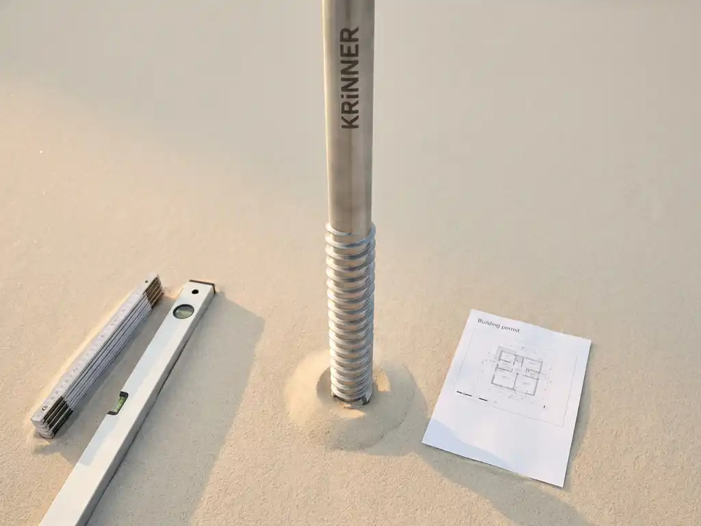 Krinner schroefpaal in wit zand met meetgereedschap en bouwvergunning, bovenaanzicht met minimalistisch ontwerp