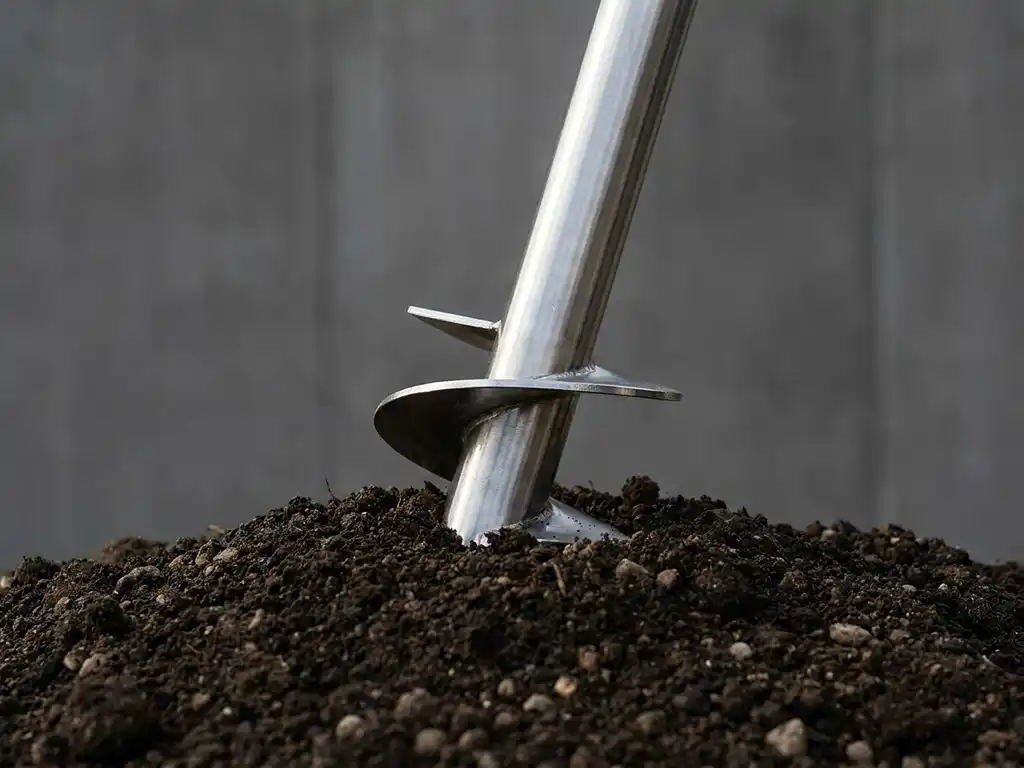 schroefpaal-staal-fundatie-grond-installatie.jpg Stalen schroefpaal met spiraaldraden steekt diagonaal uit donkere aarde met kiezels, zachte belichting op minimalistische achtergrond