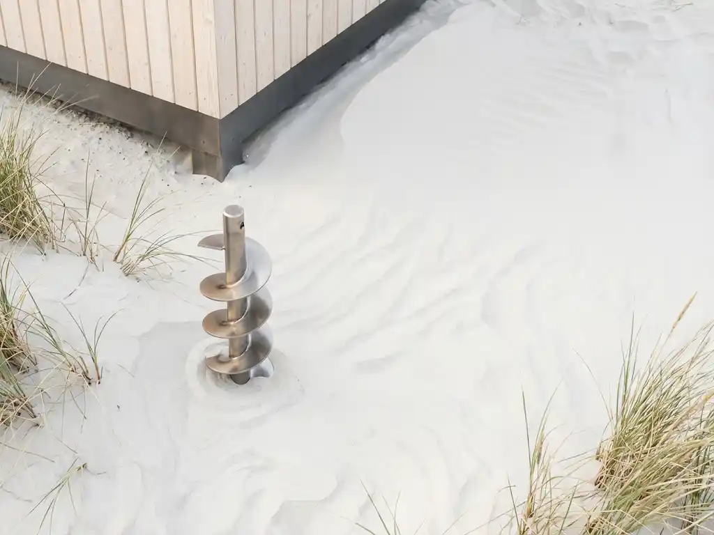 Stalen schroeffundatie spiraal in wit zand naast moderne minimalistische vakantiecabine met zachte natuurlijke belichting