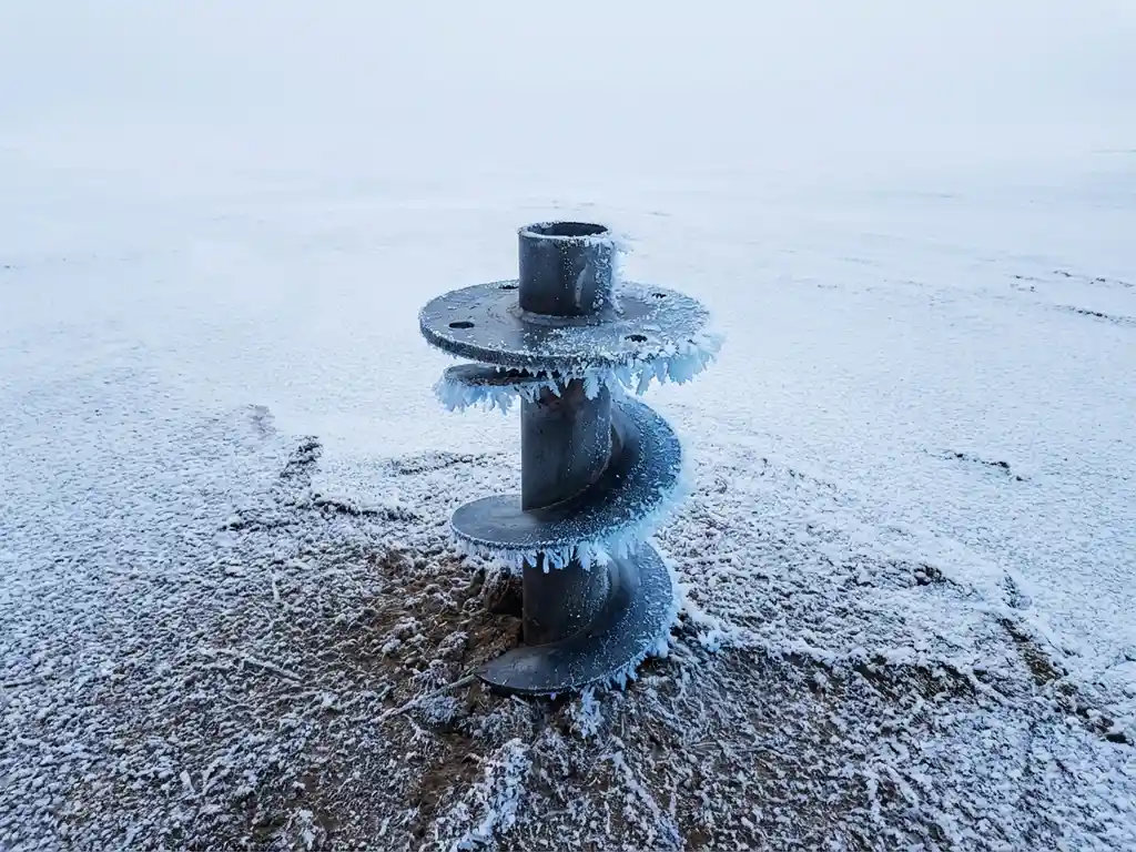 Stalen schroeffundatie paal in bevroren grond met ijskristallen aan de basis, minimalistische winterlandschap