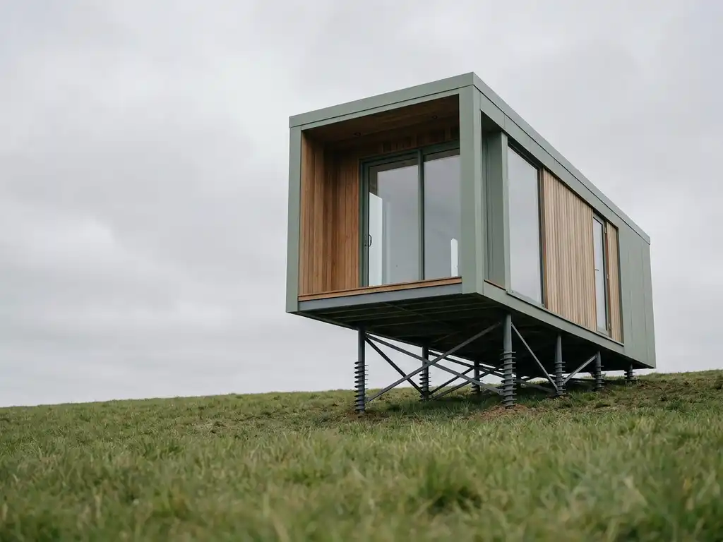 tiny-house-schroeffunderingen-helling-moderne-architectuur.jpg Kleine moderne tiny house met grote ramen op grasachtige helling, ondersteund door elegante metalen schroeffunderingen
